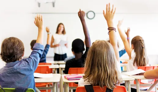 Imagem de alunos, sentados numa sala de aula, com o braço no ar para responderem à professora.