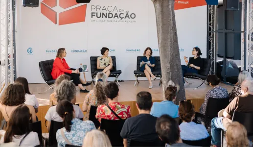 Uma plateia a assistir a um debate com quatro oradores em palco. Ao fundo, o texto "Praça da Fundação".