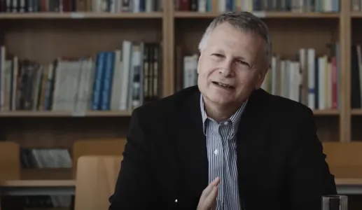 O economista político Dani Rodrik, de cabelo grisalho, sorrindo, sentado à mesa com prateleira de livros ao fundo.