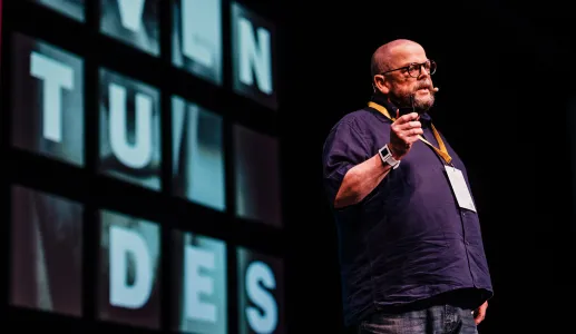 O sociólogo John Goodwin a falar em palco, a usar uma camisola escura e óculos. Texto visível: "juventudes".