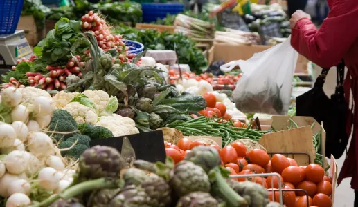 Imagem de mulher a comprar vegetais numa praça. Crédito: Canva