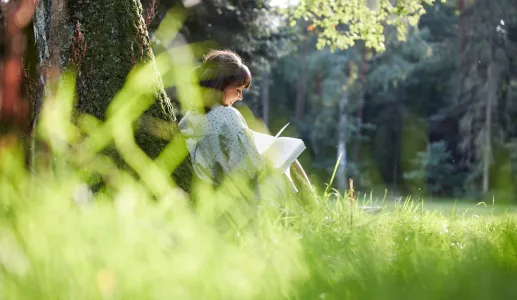 Imagem de uma rapariga a ler um livro, sentada na relva, debaixo de uma árvore. Crédito: Shutterstock