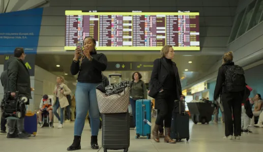 Mulher a olhar para o telemóvel em frente a um painel de chegadas e partidas do aeroporto de Lisboa.