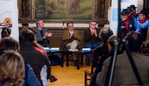 Evento com público a aplaudir, três oradores em destaque em frente a um quadro artístico.