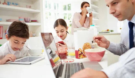 Uma família toma o pequeno-almoço na cozinha, mas o pai e os dois filhos estão a ver ecrãs de computador ou telemóvel. Crédito: Canva