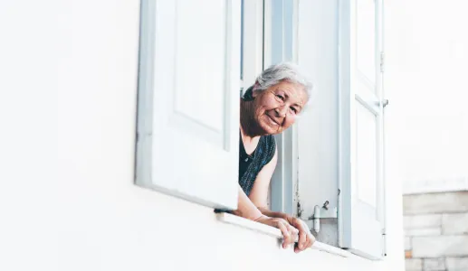 Uma mulher idosa e sorridente está à janela a espreitar para a rua, num dia de verão. Crédito: Mike Jones (Canva) 