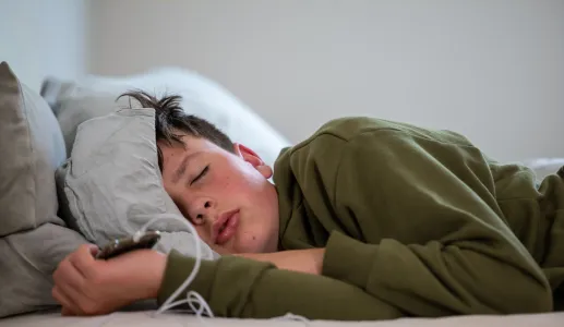 um adolescente dorme com o telemóvel na mão, sob um fundo neutro. Crédito: Canva.