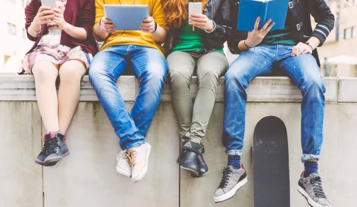 vários adolescentes estão a ver conteúdos no telemóvel e ipad, sentados num muro, num ambiente citadino. Crédito: Canva