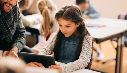 Uma menina de cabelos escuros, com um tablet na mão, tendo como fundo uma sala de aula. Crédito: Shutterstock