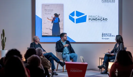 Uma plateia, de costas, a olhar para um palco, onde estão dois homens sentados a falar.