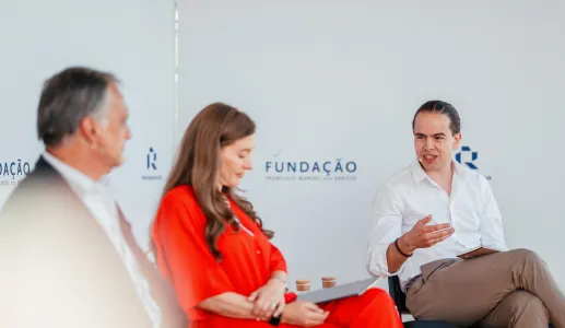 Três pessoas sentadas num palco. Dois homens de fato, uma senhora com um vestido vermelho. Ao fundo, o logotipo da Fundação.