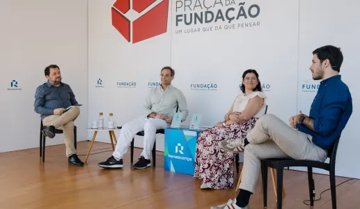 Quatro pessoas sentadas num palco, em cadeiras pretas, a apresentar um livro que está pousado em cima de uma mesa. Fundo com logotipos da Fundação.