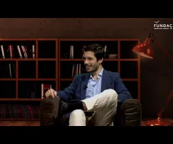Homem sentado num sofá, com uma estante de livros ao fundo e uma lâmpada acesa. Logo da Fundação no canto superior direito.