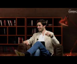 Homem de blazer claro sentado em sofá, com estante de livros ao fundo e luz vermelha. Logo da Fundação no canto superior direito.