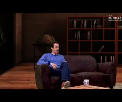 Homem sentado num sofá a falar, com um globo ao fundo e estantes de livros. Texto: "FUNDAÇÃO".