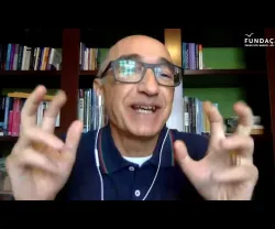 Homem com óculos, gesticulando em frente a uma estante de livros. Logotipo da Fundação.