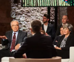 Discussão em painel com duas pessoas a falar e público a ouvir, luz suave ao fundo.