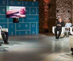 Debate com várias pessoas sentadas, tela ao fundo com duas mãos entrelaçadas."FRONTEIRAS XXI".