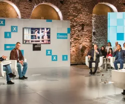 Imagem de um painel de discussão com várias pessoas sentadas em cadeiras brancas.