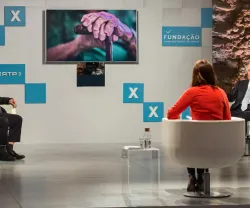Imagem de um debate com quatro pessoas sentadas e uma imagem de mão idosa numa tela, ao fundo.