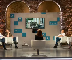 Painel de discussão com várias pessoas sentadas em cadeiras brancas, num estúdio de televisão.