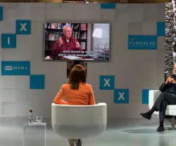 Mesa redonda com quatro participantes, ecrã a mostrar um orador e legendas.