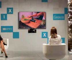 Três mulheres em cadeiras brancas, com um ecrã a mostrar vegetais a serem preparados.