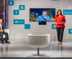 Apresentadora em estúdio com painel, exibição de vídeo e logos da RTP e Fundação.