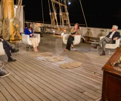 Grupo de pessoas a falar, sentadas num navio, à noite. Textos não visíveis.