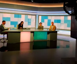Estúdio de notícias com apresentadores sentados à mesa e fundo moderno em tons de azul.