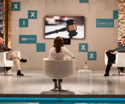 Debate com três pessoas sentadas em cadeiras; um martelo de juiz na tela atrás deles.