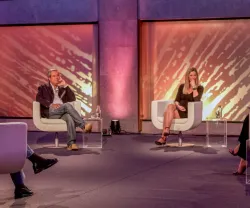 Painel de debate com quatro pessoas sentadas em cadeiras brancas. Fundo colorido.