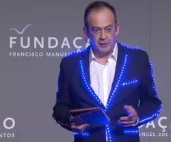 Homem de fato delineado com luzes, em palco, a apresentar um evento da Fundação.
