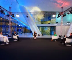 Painel de discussão com cinco pessoas, sentadas em cadeiras brancas, num ambiente moderno e iluminado.