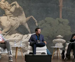 Três pessoas sentadas em cadeiras, a discutir em frente a um fundo artístico.