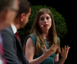 Mulher a falar num painel, a gesticular, e com um microfone na orelha. Fundo com plantas.