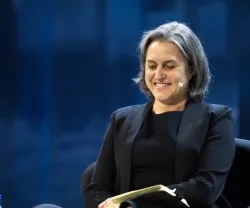 Mulher sorridente sentada em palco, segura um caderno. Luzes azuis ao fundo.
