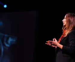 Mulher de perfil, a falar no palco, com fundo escuro. 