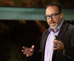 O fundador da Wikipedia Jimmy Wales a gesticular enquanto discursa em palco, sobre um fundo escuro.