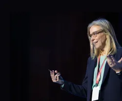 Mulher de cabelo loiro e óculos faz uma apresentação em palco, gesticulando com uma caneta na mão.