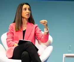Mulher com blazer rosa fala, enquanto segura um caderno e gesticula. Fundo azul.