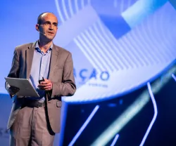 Carlos Jalali, professor de Ciência Política, em pé, a discursar, enquanto segura um tablet na mão.