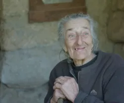 Retrato de uma mulher idosa, com as mãos apoiadas no cimo do cabo de uma vassoura. Atrás, uma parede de pedra.