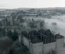 Castelo envolto em névoa, com vista para uma cidade nas colinas ao fundo.