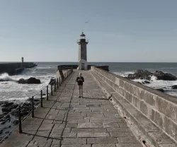 Homem a caminhar num caminho de pedra, rumo a um farol, junto ao mar agitado.