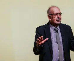 Um homem de óculos e fato, a gesticular durante uma apresentação. Fundo amarelo