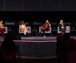 Três pessoas sentadas em sofás num palco. Um homem fala enquanto as outras escutam.