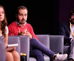 Três pessoas sentadas em sofás num palco. Um homem fala enquanto as outras escutam.