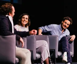 Os humoristas Eduardo Madeira, Bumba na Fofinha e Carlos Coutinho Vilhena sorridentes, sentados em cadeiras. Fundo preto.