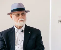 O biblófilo Alberto Manguel sentado, com um chapéu na cabeça e de óculos. Um microfone de lapela preso na camisa.
